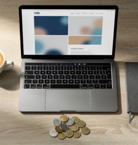 Laptop showing a simple website beside a coffee, a notebook and a small pile of coins on a wooden desk