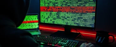 Hooded figure at a keyboard, monitor and laptop glowing with green terminal code, a line highlighted in red signifying an exploit